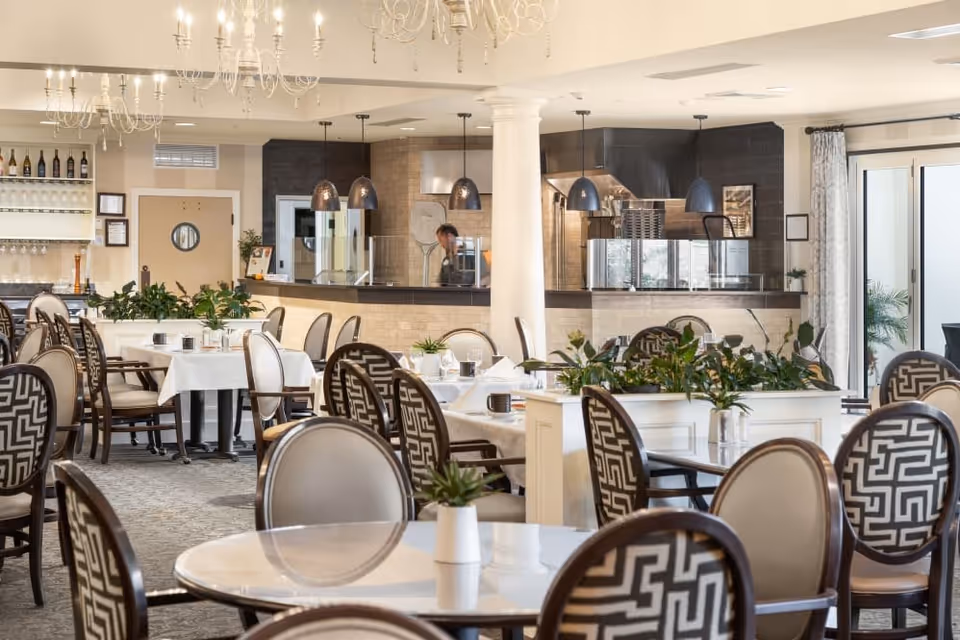 A bright and elegant dining room in an independent living community featuring round and rectangular tables with white tablecloths, patterned chairs, and small potted plants. The background shows a kitchen area with pendant lights and a person working behind a counter.