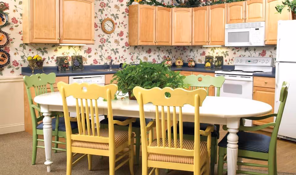 A bright kitchen with light wood cabinets, a white refrigerator, stove, and microwave. A white oval dining table is surrounded by six chairs painted in yellow and green. The walls are decorated with floral wallpaper, and a green leafy plant is placed in the center of the table.