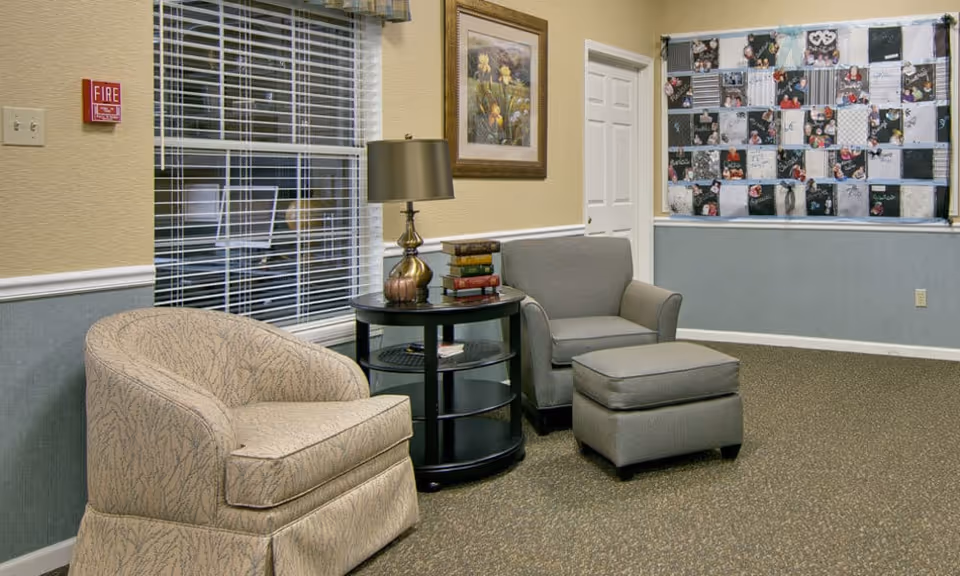 Small seating area with two upholstered chairs, an ottoman, a round side table with a lamp and stacked books, and a bulletin board on the wall.