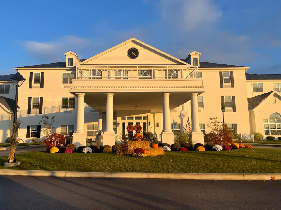 Front exterior view of a senior living facility named Apple Blossom Senior Living, featuring a large white building with columns at the entrance, a clock above the entrance, and fall-themed decorations including pumpkins and hay bales on the lawn.