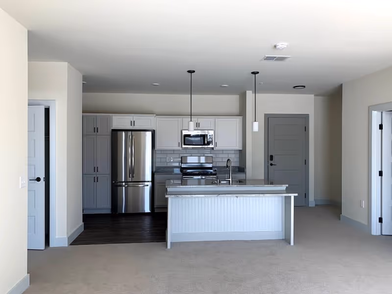 Open-plan interior showing a modern kitchen with a central island, stainless steel refrigerator and stove, pendant lights, and adjoining living area.