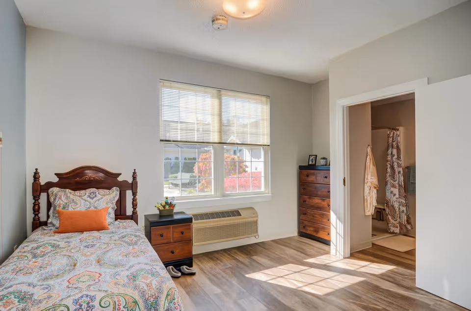 A bright senior living bedroom with a single wooden bed featuring a colorful patterned bedspread and an orange pillow. Next to the bed is a small wooden nightstand with a plant on top. A large window with blinds lets in natural light, illuminating the room. Across from the bed is a tall wooden dresser. An open door reveals a bathroom with a shower curtain and bathrobe hanging on the wall. The room has light-colored walls and wood flooring.