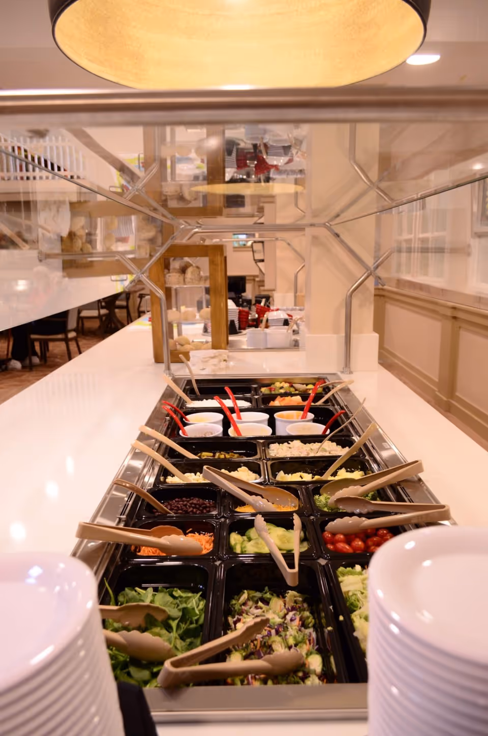 A salad bar with various fresh vegetables and toppings including lettuce, cherry tomatoes, cucumbers, shredded carrots, black beans, and several dressings in cups. There are tongs and serving spoons placed in each container, and stacks of white plates are visible at the front. The background shows a dining area with tables and chairs.