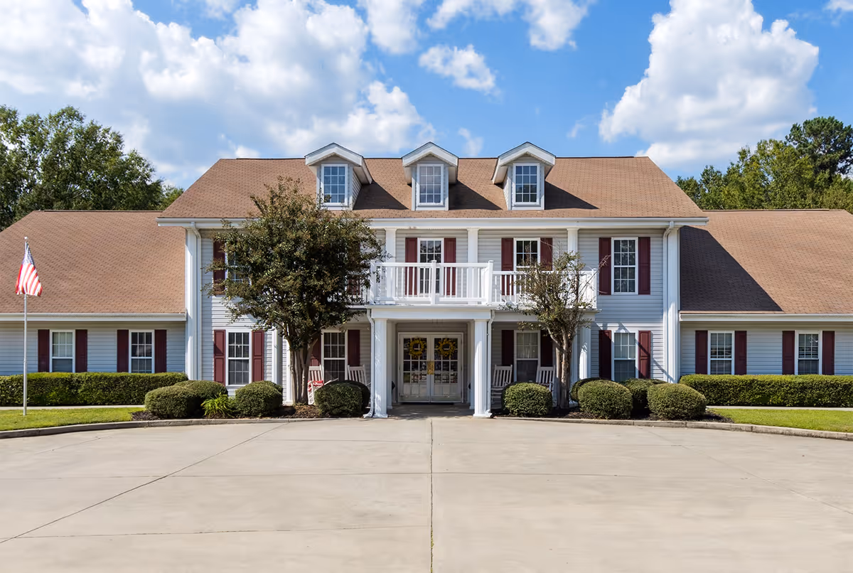 Front exterior view of Hitchcock Square facility, a two-story building with white siding, brown roof, red shutters, and a balcony above the entrance. There are two trees and bushes in front, an American flag on the left, and a clear blue sky with some clouds.
