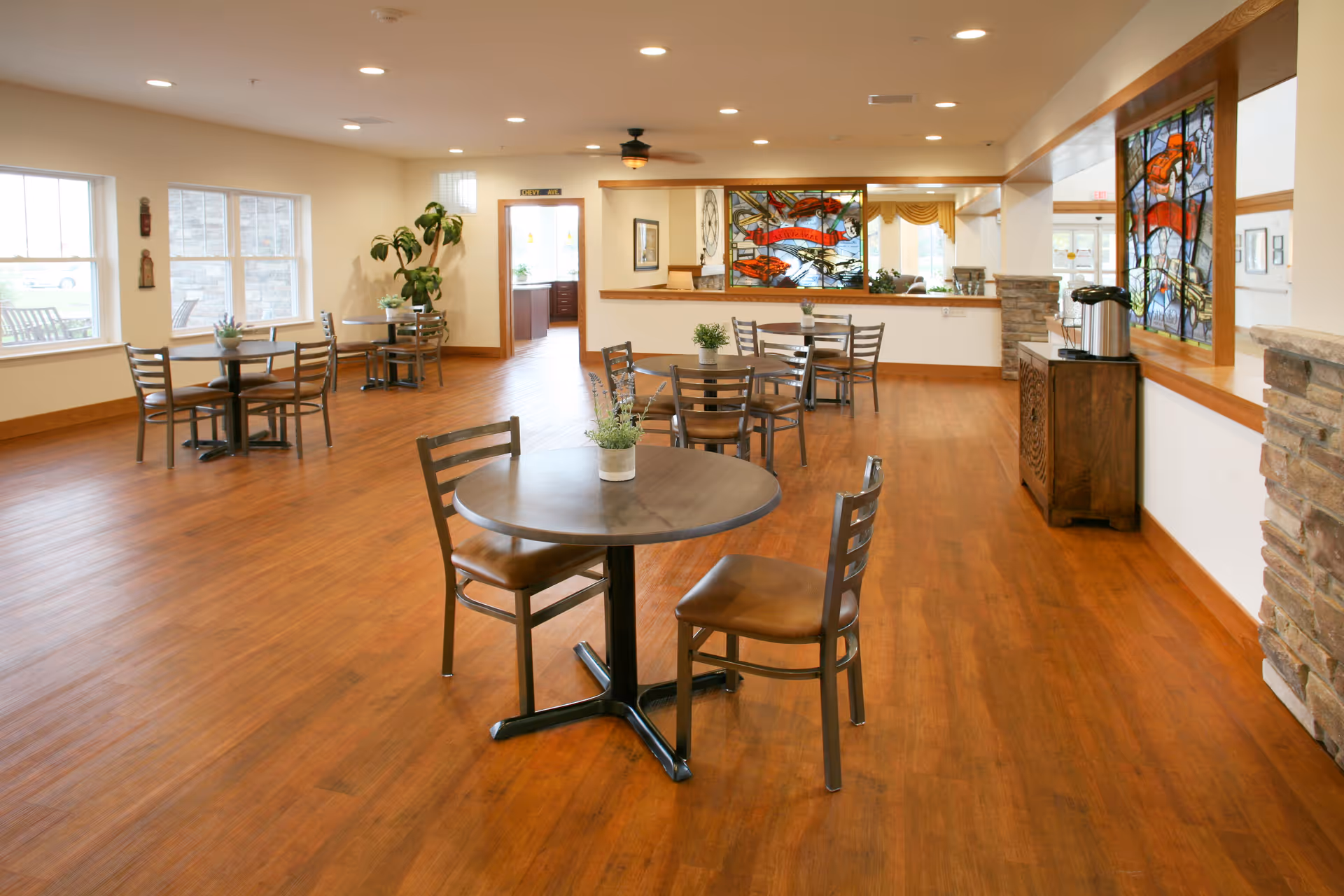 Spacious dining/common area with round tables and chairs on wood floors, stained-glass panels and large windows.