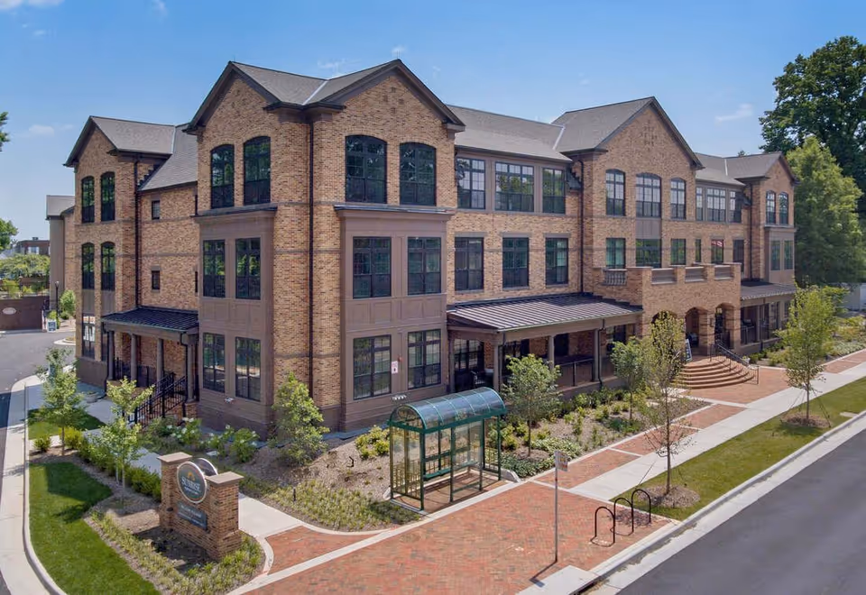Exterior view of a large three-story brick building with multiple windows, a covered entrance, and landscaped surroundings including small trees and shrubs. A bus stop shelter is visible near the sidewalk in front of the building under a clear blue sky.