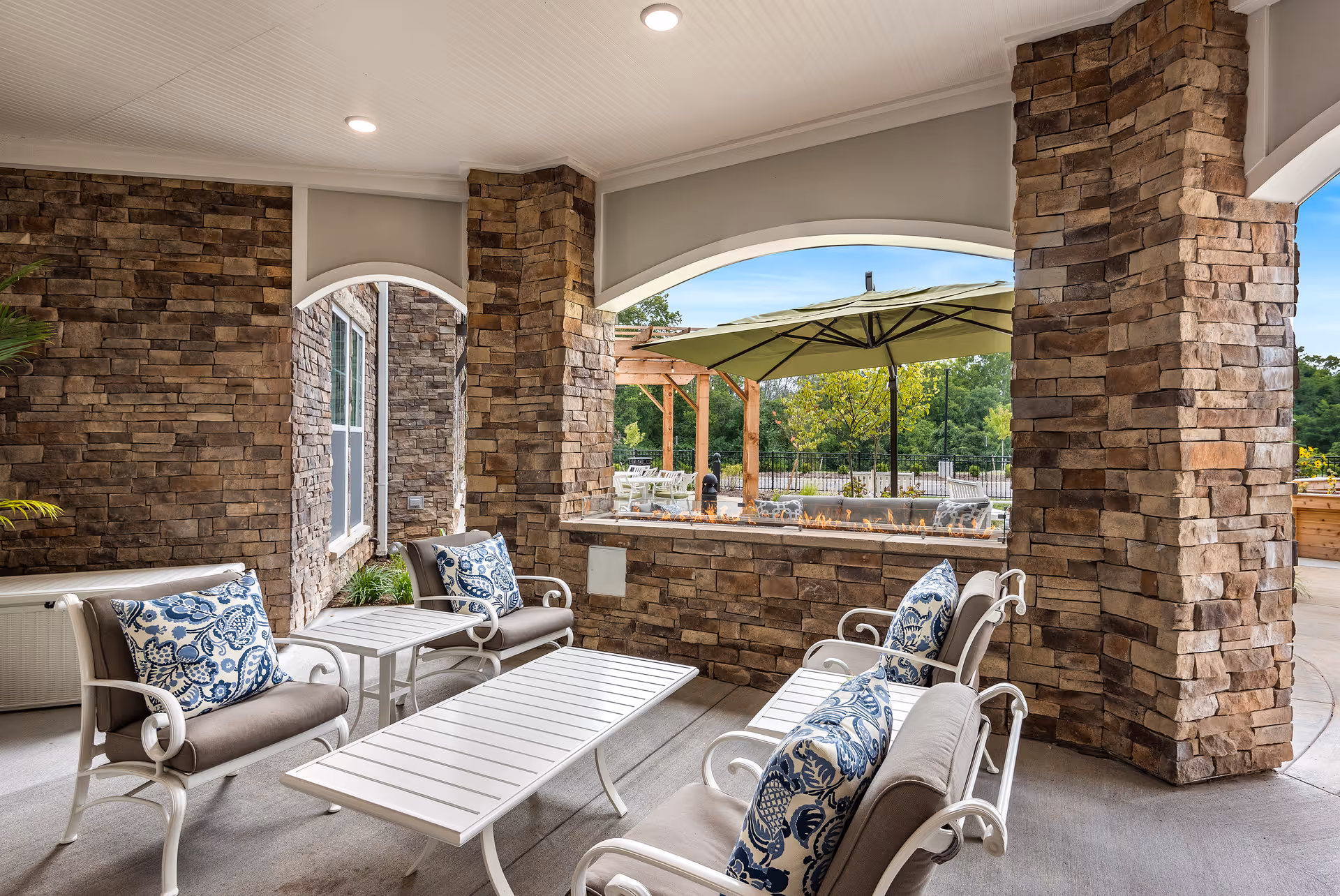 Covered outdoor seating area with stone pillars and walls, featuring white metal chairs with beige cushions and blue patterned pillows, white tables, and a large green umbrella visible through an arched opening overlooking a garden area.