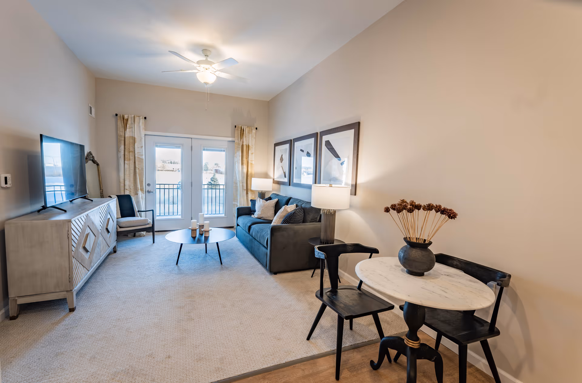 A bright living room with beige walls and carpeted floor featuring a gray sofa with cushions, a round coffee table with candles, a TV on a wooden console, two black chairs around a small round marble table with a vase of dried flowers, and French doors with curtains leading to a balcony.