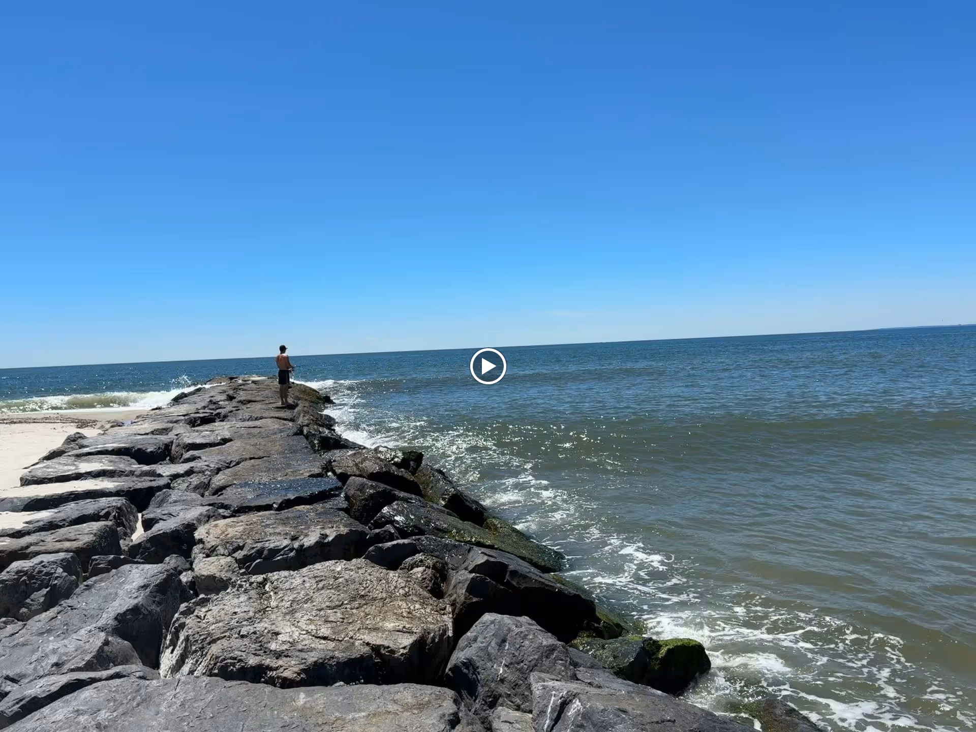 A person standing on a rocky jetty extending into the ocean under a clear blue sky with waves crashing against the rocks.