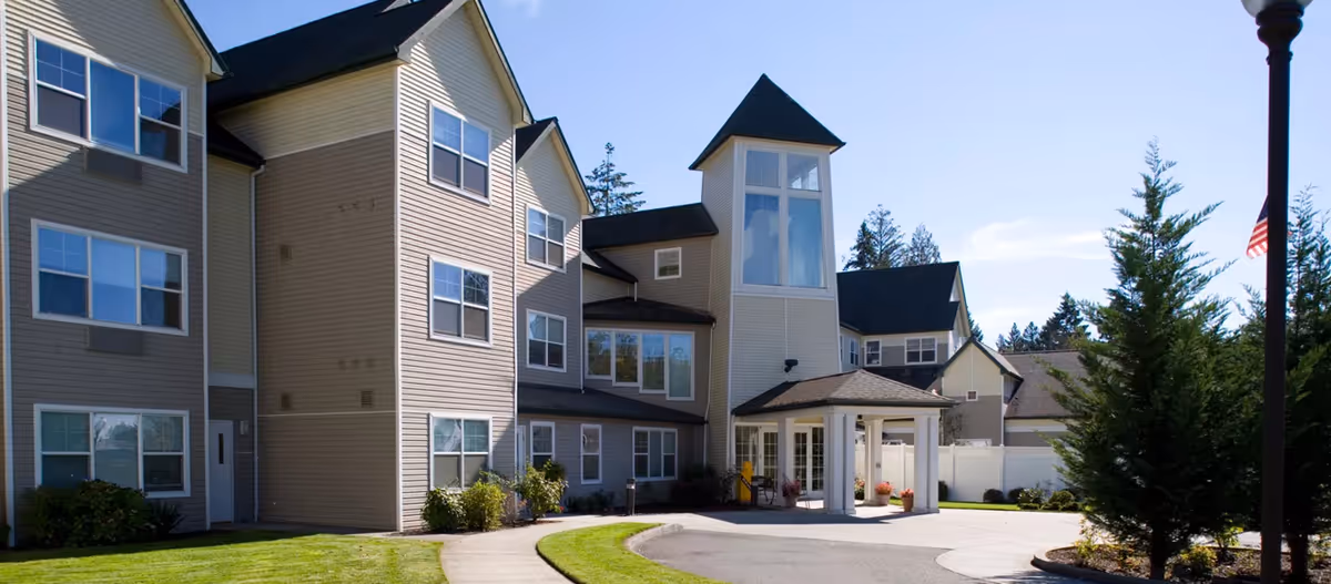 Front exterior of a multi-story senior living building with a covered entrance, landscaped lawn, and driveway under a clear sky.