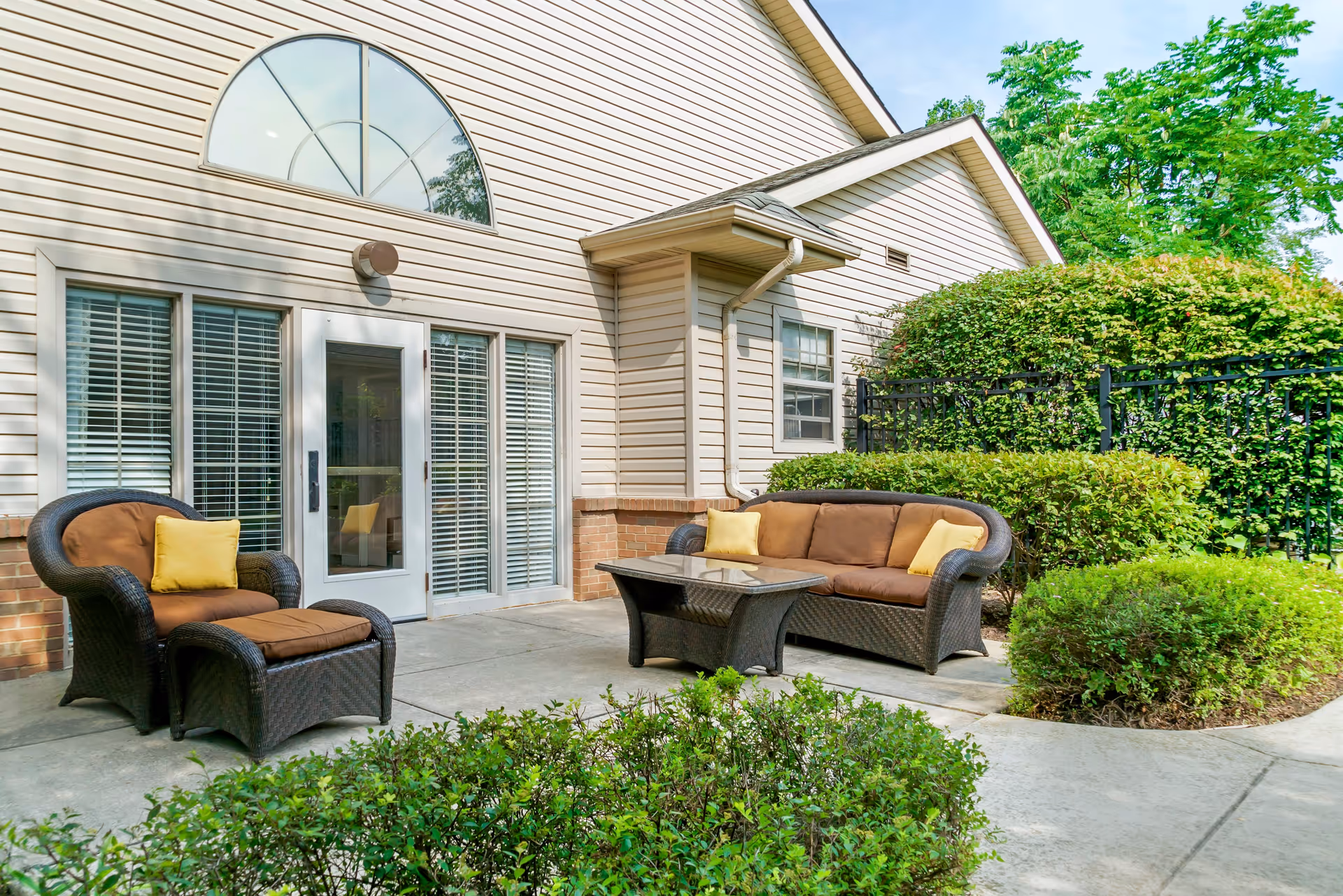 Outdoor patio area at Addington Place of Northville featuring wicker furniture with brown cushions and yellow pillows, a glass-top coffee table, surrounded by green bushes and trees, adjacent to a beige building with large windows and a glass door.