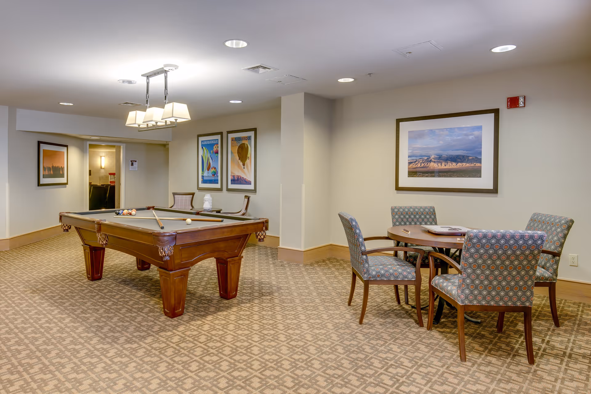A recreational room featuring a pool table with cues and balls set up, a round table with four patterned chairs, and framed artwork on the walls. The room has carpeted flooring and recessed ceiling lights.