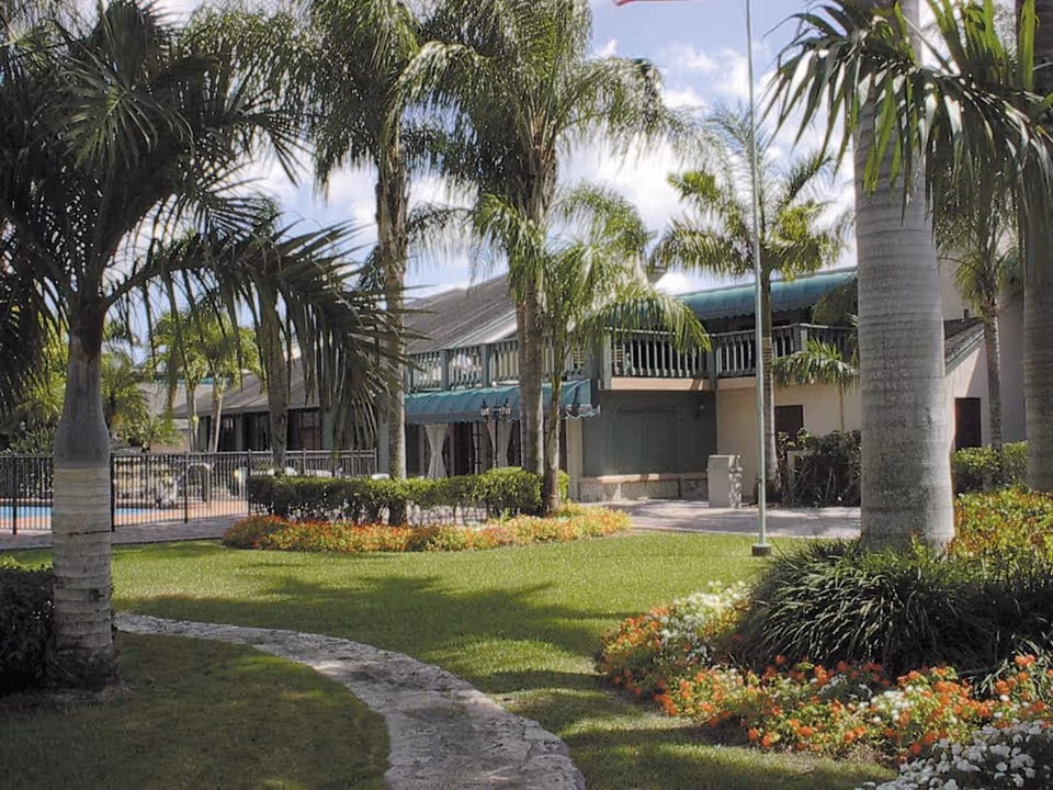 Well-manicured courtyard with palm trees, flower beds, a curved stone walkway, and a two-story building with balconies and a fenced pool area.
