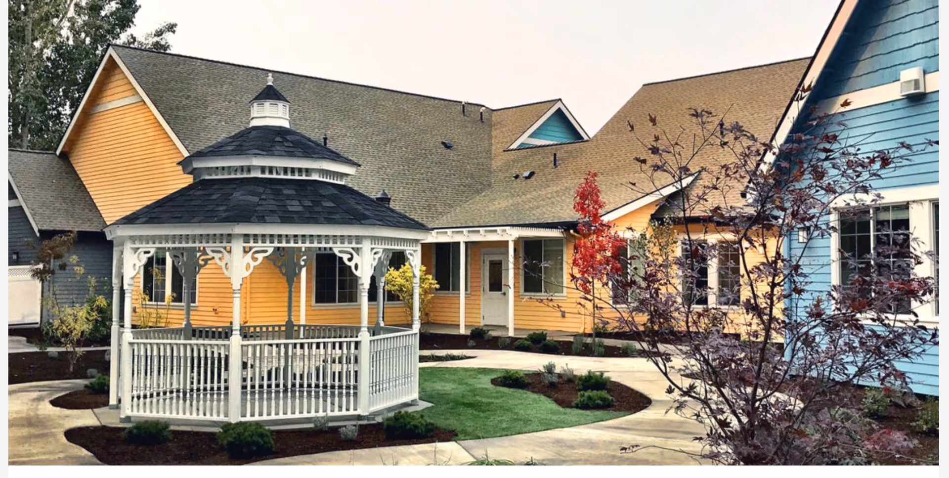 Outdoor courtyard area of a senior living facility with a white gazebo in the center, surrounded by a paved walkway, small landscaped bushes, and colorful buildings painted in yellow and blue.