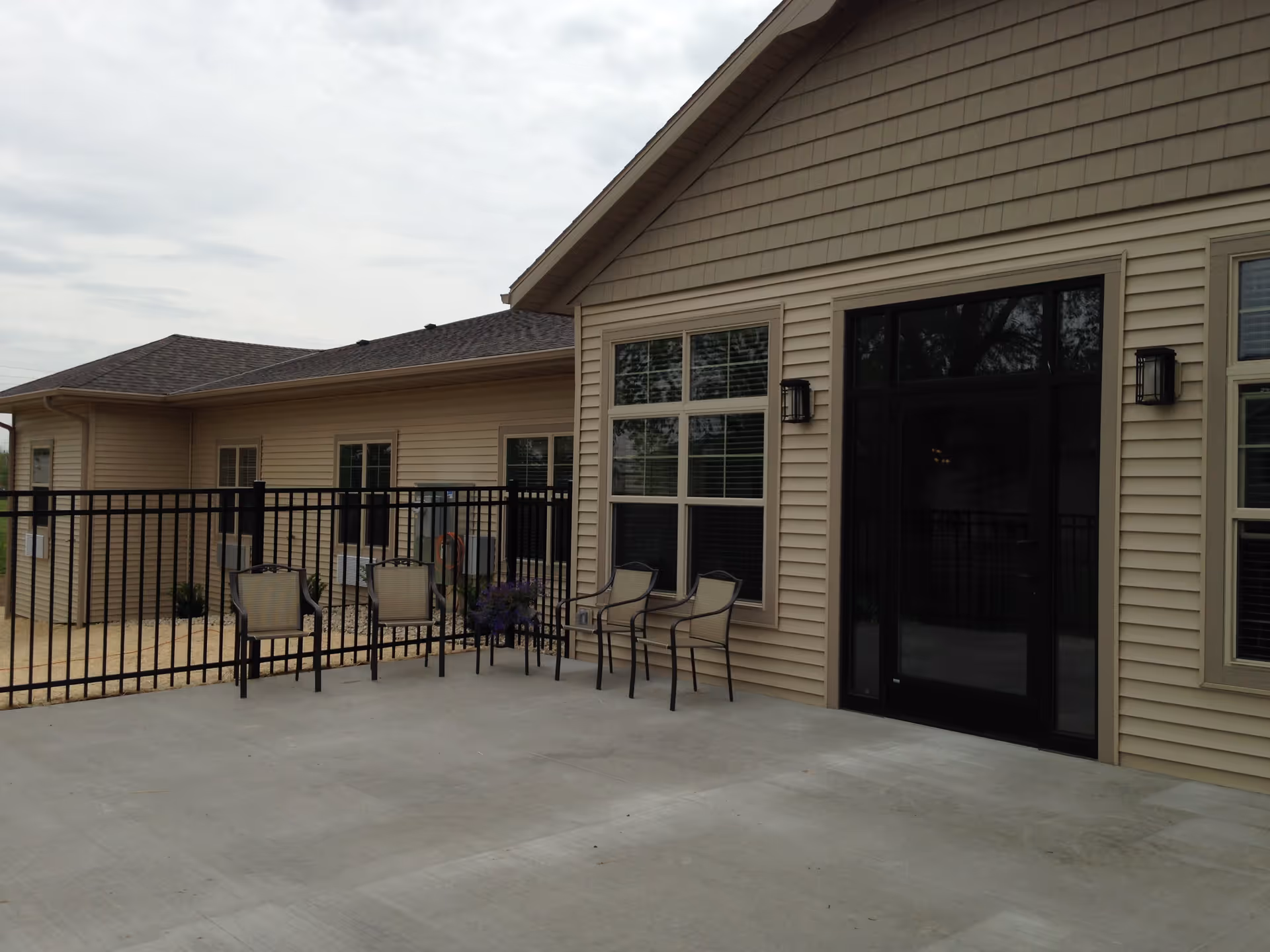 Outdoor patio area at Lilac Springs Assisted Living Center with four chairs arranged near a black metal fence and beige siding building with windows and a black door.