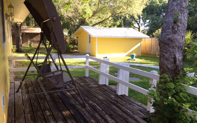 A wooden deck with a brown swing chair attached to a metal frame, surrounded by white railings. In the background, there is a yellow shed with a white roof, green grass, trees, and a wooden fence.