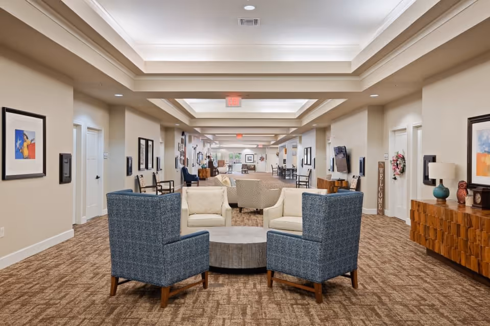 Spacious, well-lit senior living lounge with armchairs and sofas arranged around a round table in a long carpeted corridor.