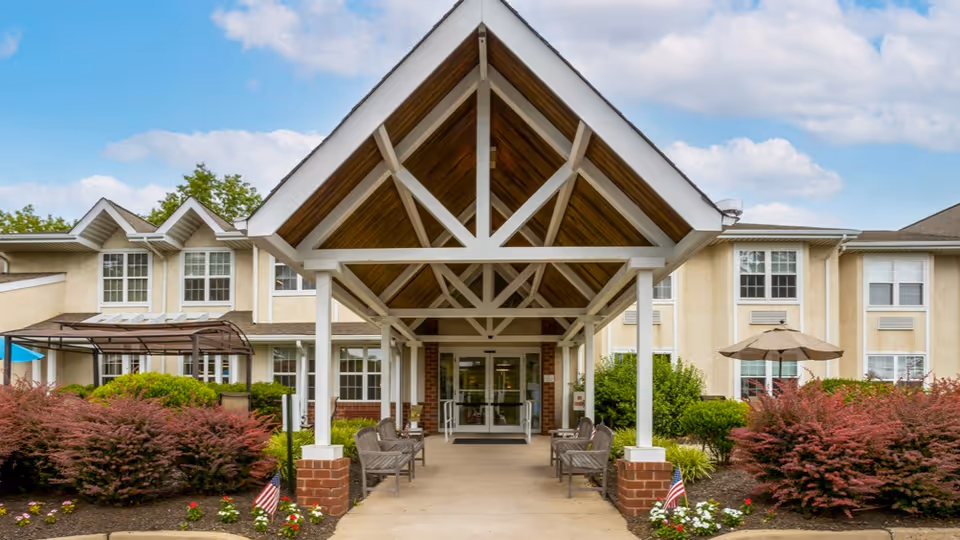 Front exterior view of Brookdale Northampton senior living facility showing a covered entrance with white pillars and wooden ceiling beams, benches on either side of the walkway, landscaped bushes and flowers, and a clear blue sky with some clouds.