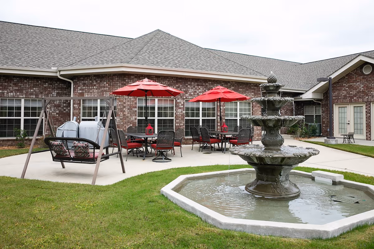 Outdoor patio area at Hearthside Senior Living featuring a multi-tiered water fountain in the foreground, a swing bench with cushions, and several tables with red umbrellas and chairs on a concrete patio next to a brick building.