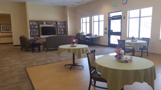 Bright common room with round tables covered in green tablecloths, chairs, and a seating area with bookshelves and a TV.