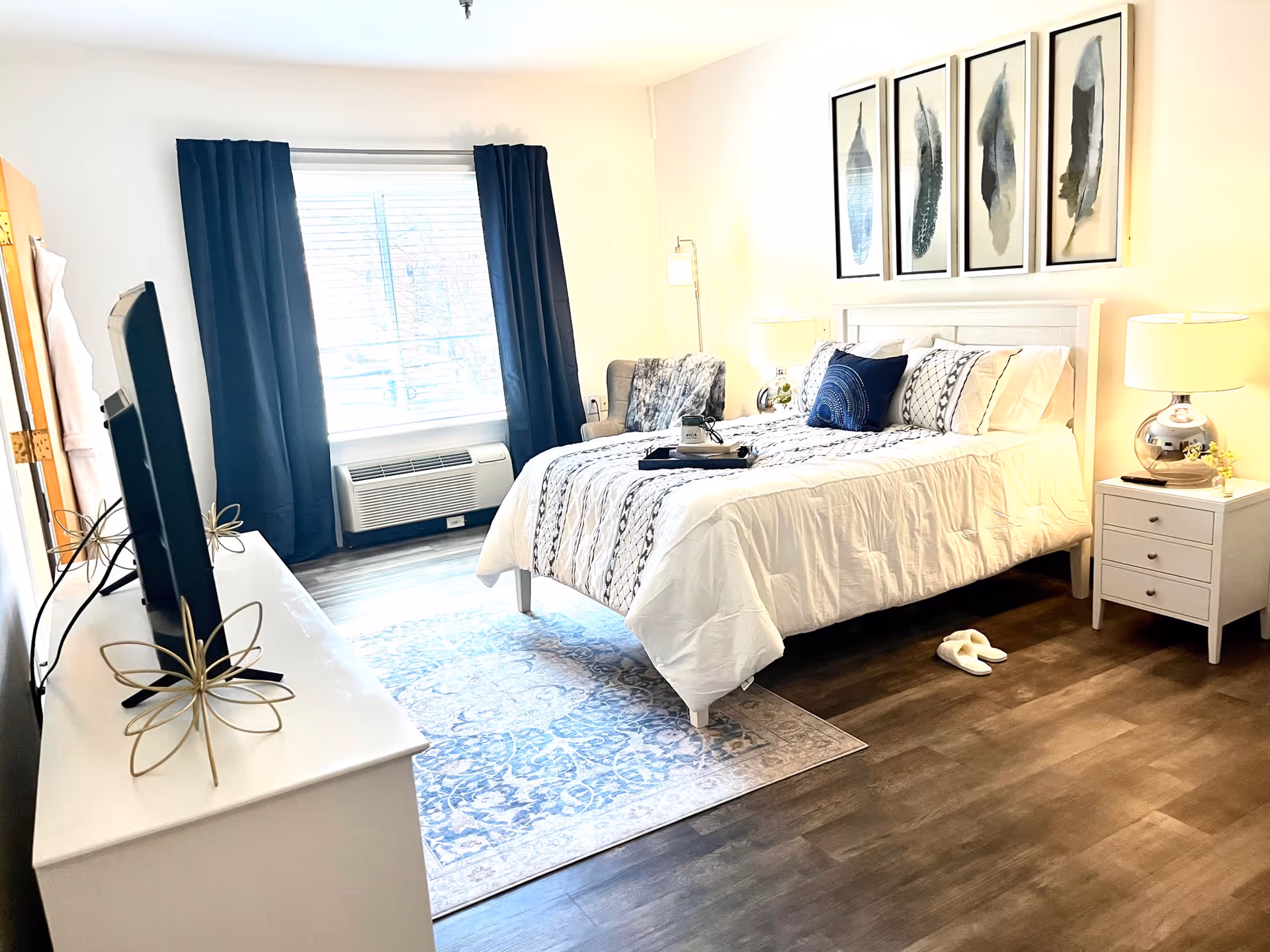 Bright furnished bedroom with a bed, nightstands, dresser with TV, armchair, and a window with dark curtains.