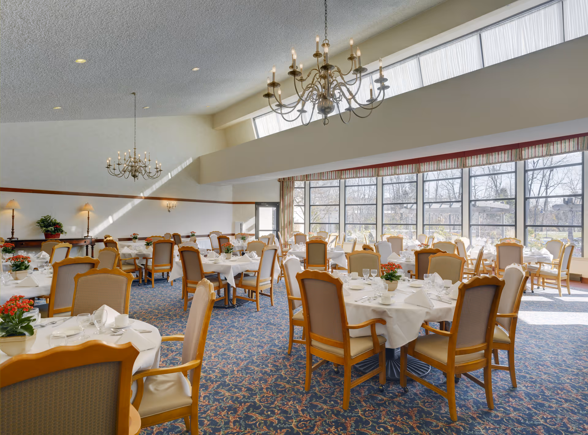 A bright and spacious dining room with multiple round tables covered in white tablecloths, each set with glassware, plates, and napkins. The room features large windows allowing natural light to fill the space, patterned carpet flooring, wooden chairs with cushioned seats, chandeliers hanging from the ceiling, and decorative plants on the tables.