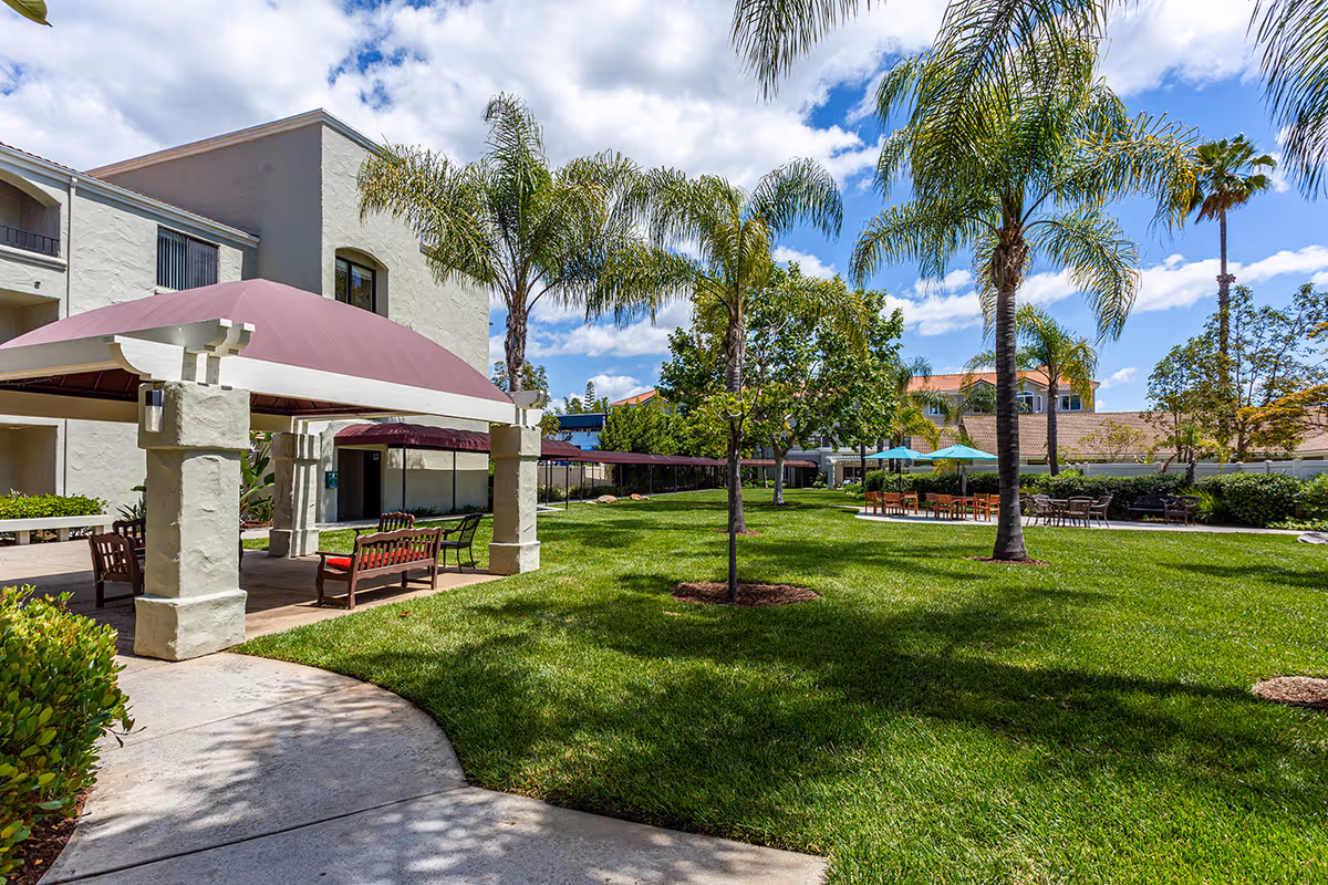 Outdoor garden area at Las Villas Del Norte featuring a covered seating area with benches, palm trees, green grass, and additional tables with umbrellas under a partly cloudy sky.