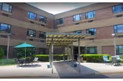 Exterior view of a three-story brick building with multiple windows, a covered entrance with a pergola structure, outdoor seating with tables and umbrellas, and some greenery along the building perimeter.