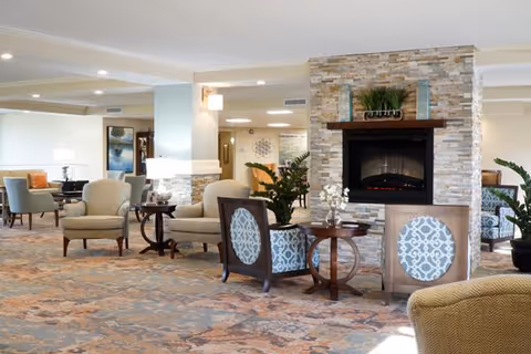 A spacious and well-lit common area in a retirement community featuring comfortable armchairs and small round tables arranged around a modern stone fireplace. The room has a patterned carpet, potted plants, and soft lighting, creating a cozy and inviting atmosphere.