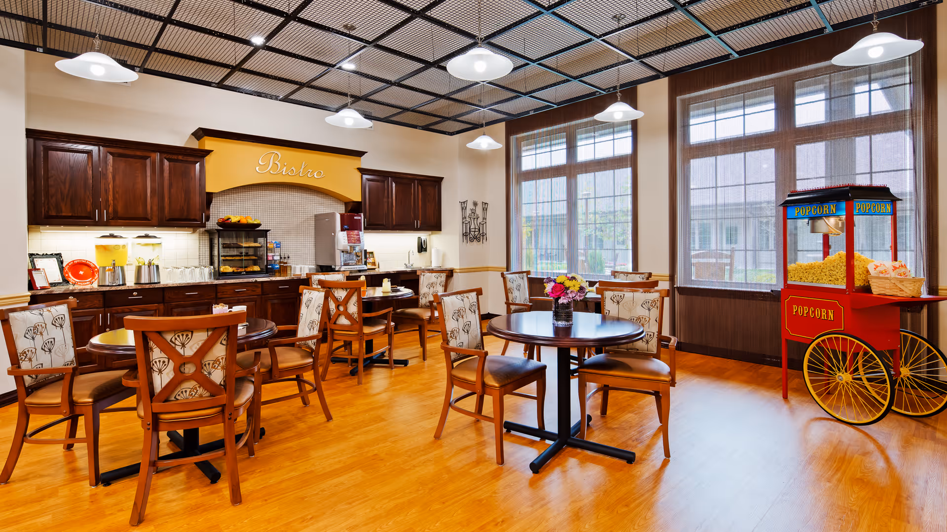 A bright and inviting dining area with wooden floors and multiple round tables surrounded by wooden chairs with patterned cushions. The back wall features dark wood cabinets and a counter with a coffee machine, juice dispensers, and a small food display under a yellow sign that reads 'Bistro'. Large windows allow natural light to fill the room, and a red popcorn machine is positioned near the windows.