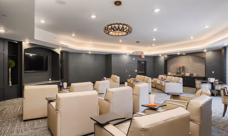 A modern lounge area with beige leather armchairs arranged in rows facing a wall-mounted flat screen TV. The room features dark walls, recessed lighting, two decorative ceiling light fixtures, and a popcorn machine on a counter in the back corner. There are also small tables with chairs near the windows on the right side.