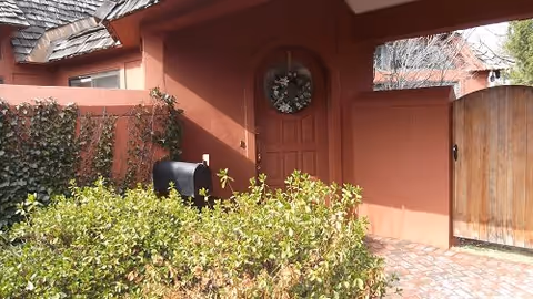 Exterior view of a residential-style building entrance with a reddish-brown stucco wall, a wooden door decorated with a wreath, a black mailbox, and surrounding greenery including bushes and ivy.