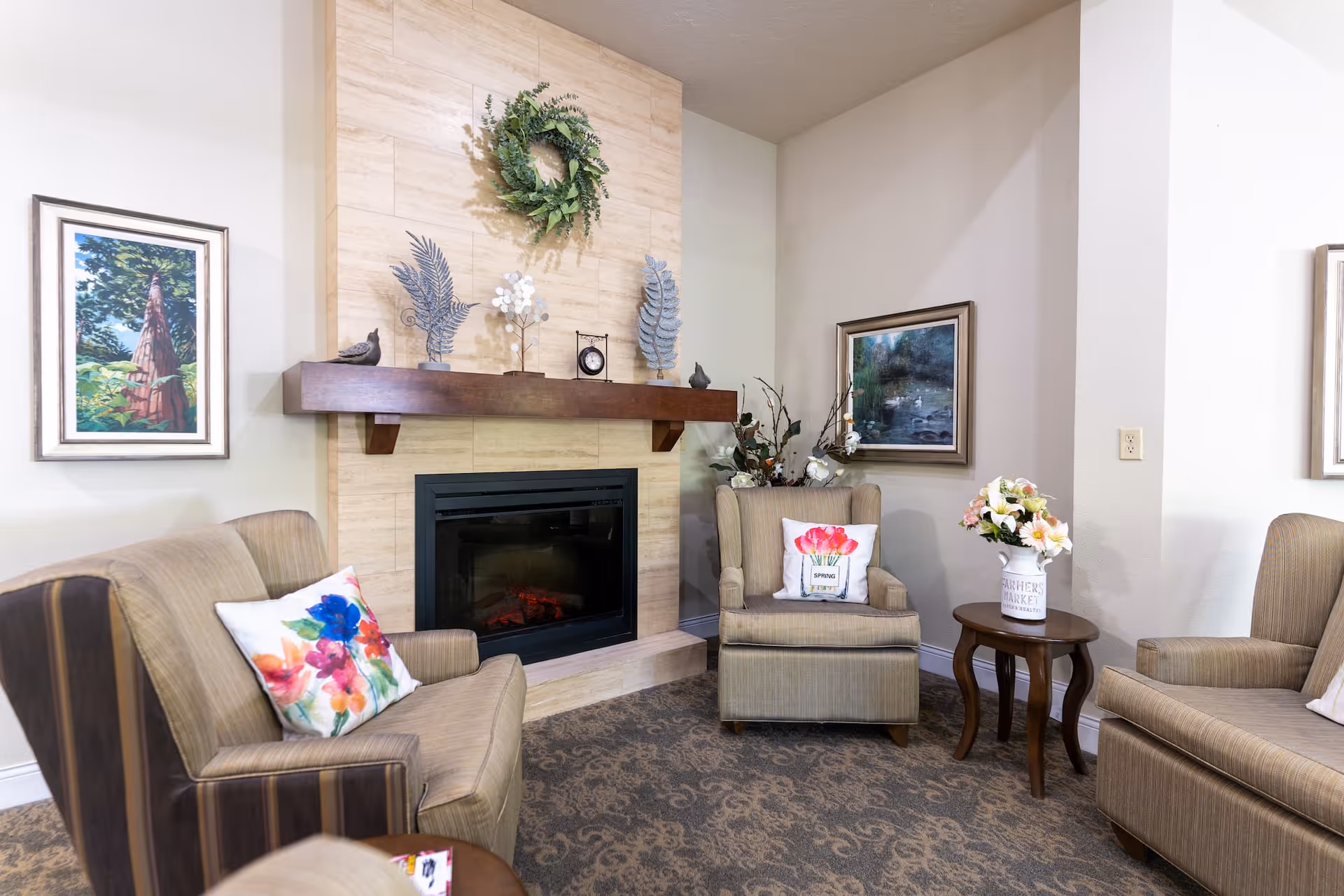Cozy common room with armchairs arranged around a fireplace, decorative mantel, and a side table with flowers.