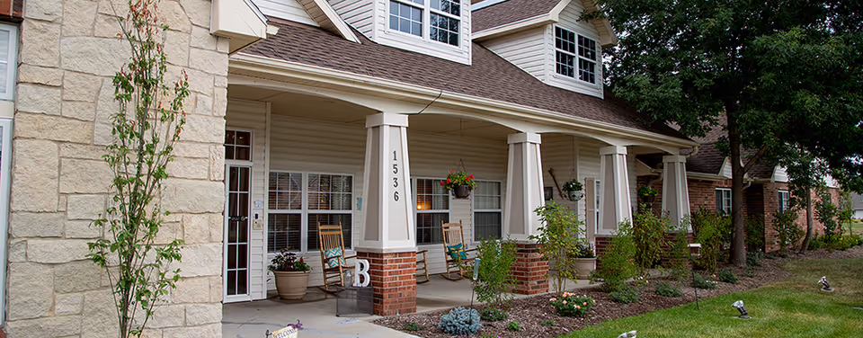 Front porch of a senior living facility with rocking chairs, hanging flower pots, and a well-maintained garden with shrubs and small trees. The building has beige siding, brick pillars, and large windows with white trim. The address number 1536 is visible on one of the pillars.