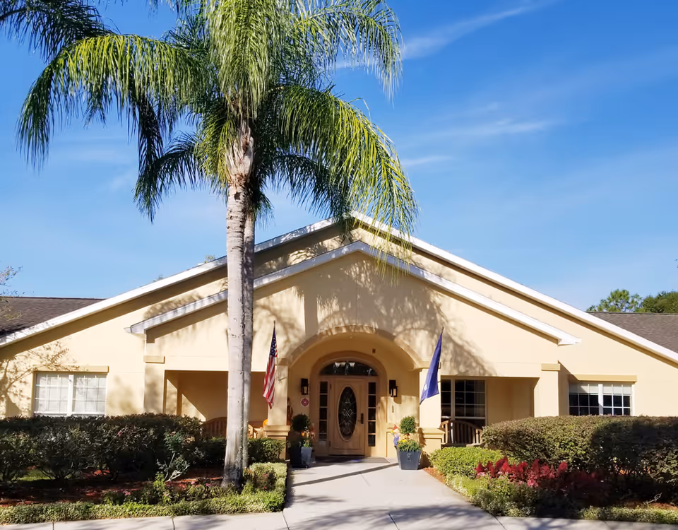 Front exterior view of a single-story building with a beige facade, an arched entrance door with glass panels, two flags on either side of the entrance, a tall palm tree in front, and well-maintained landscaping including bushes and flowers under a clear blue sky.