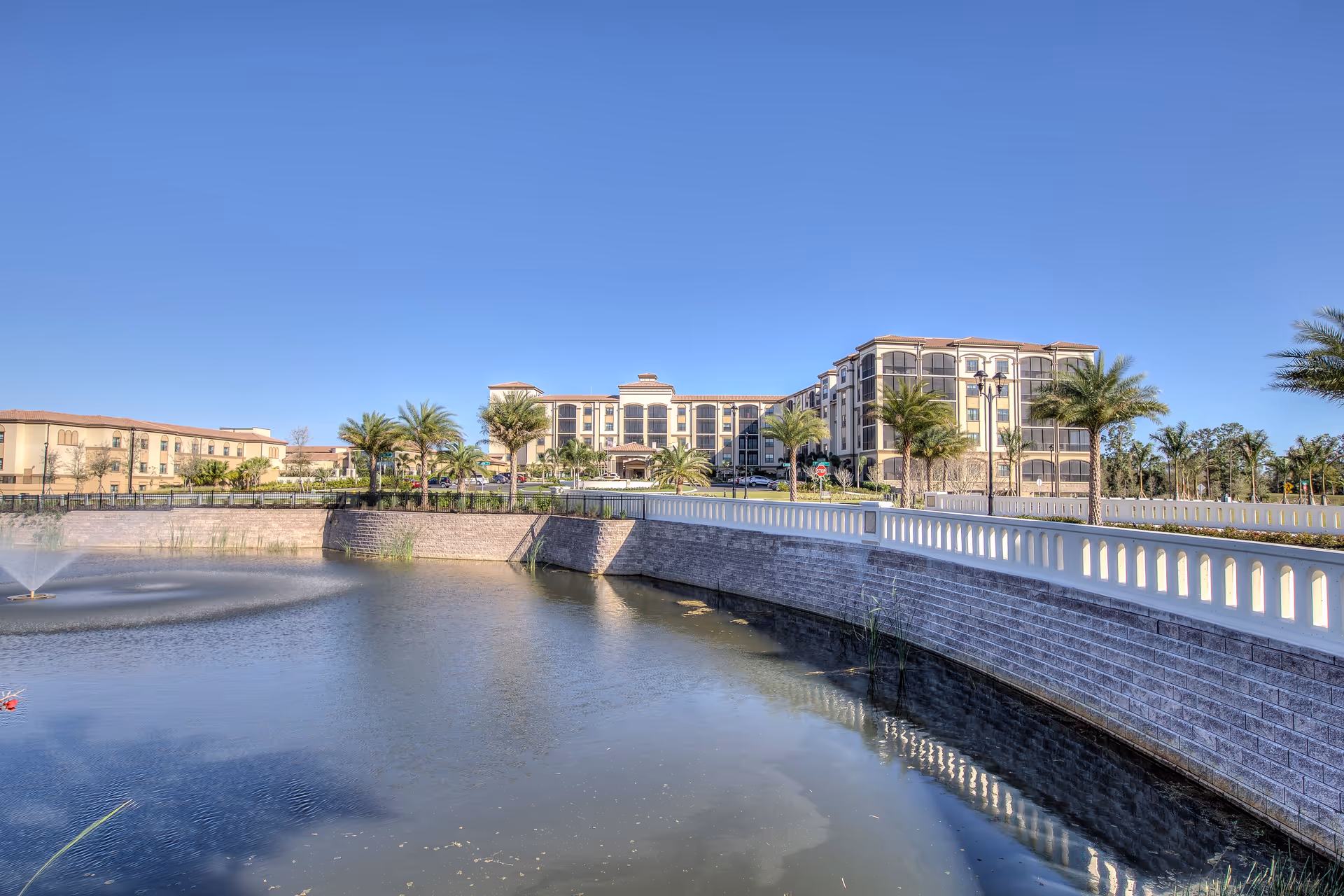 Exterior view of The Arlington of Naples senior living facility with a large pond and fountain in the foreground, palm trees lining the walkway, and a clear blue sky above.