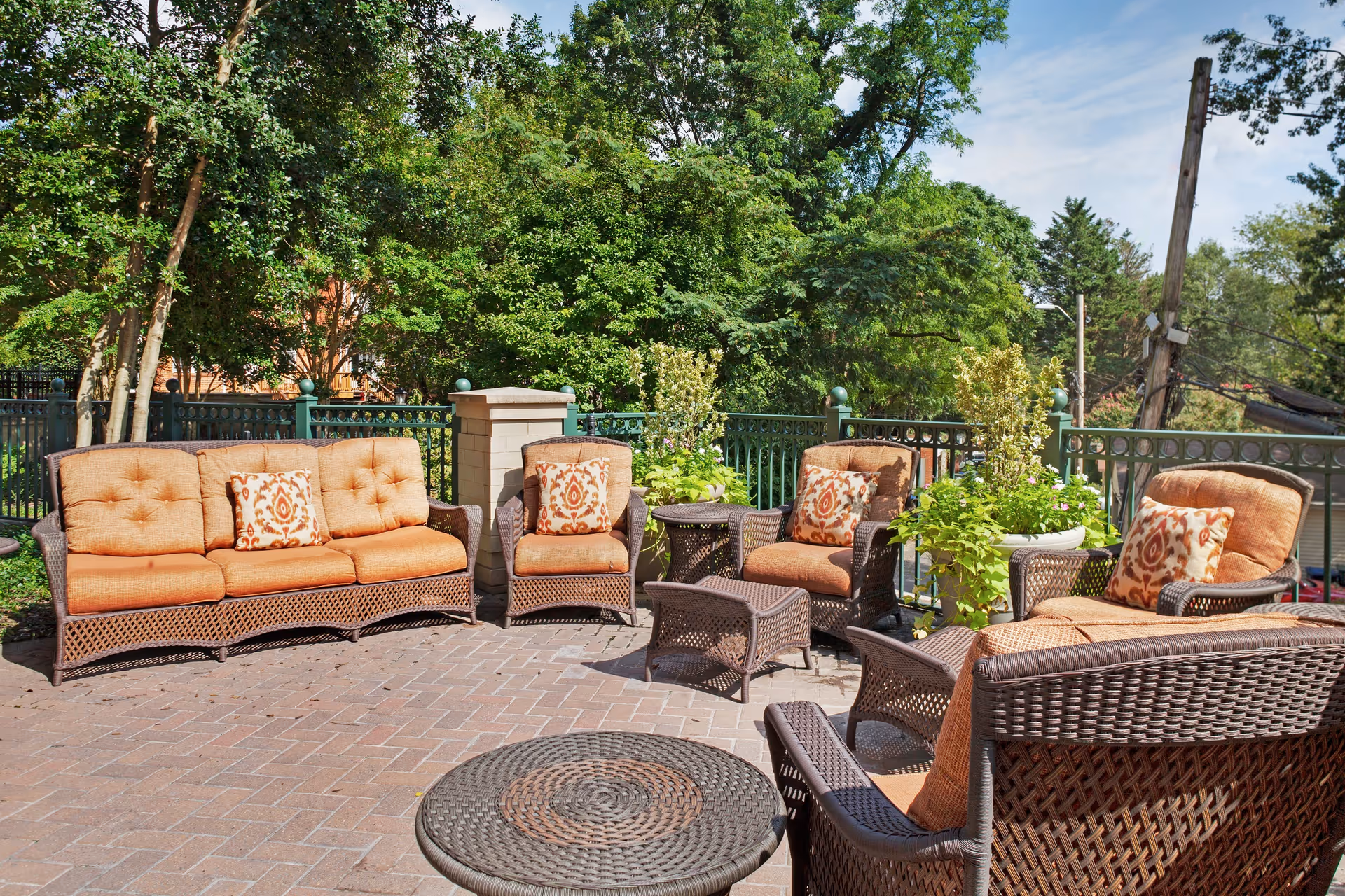 Outdoor patio with wicker seating and orange cushions, potted plants, and a brick floor under trees.