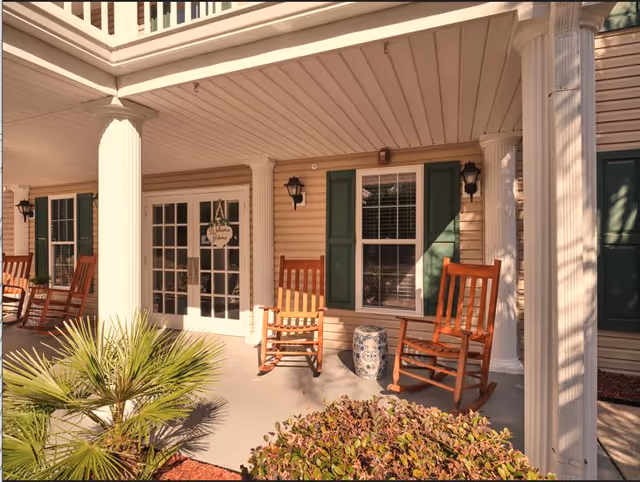 Front porch area of a building with beige siding, white columns, and green window shutters. Two wooden rocking chairs and a decorative ceramic stool are placed on the porch. A glass door with a welcome sign is visible in the background, along with outdoor wall lanterns.