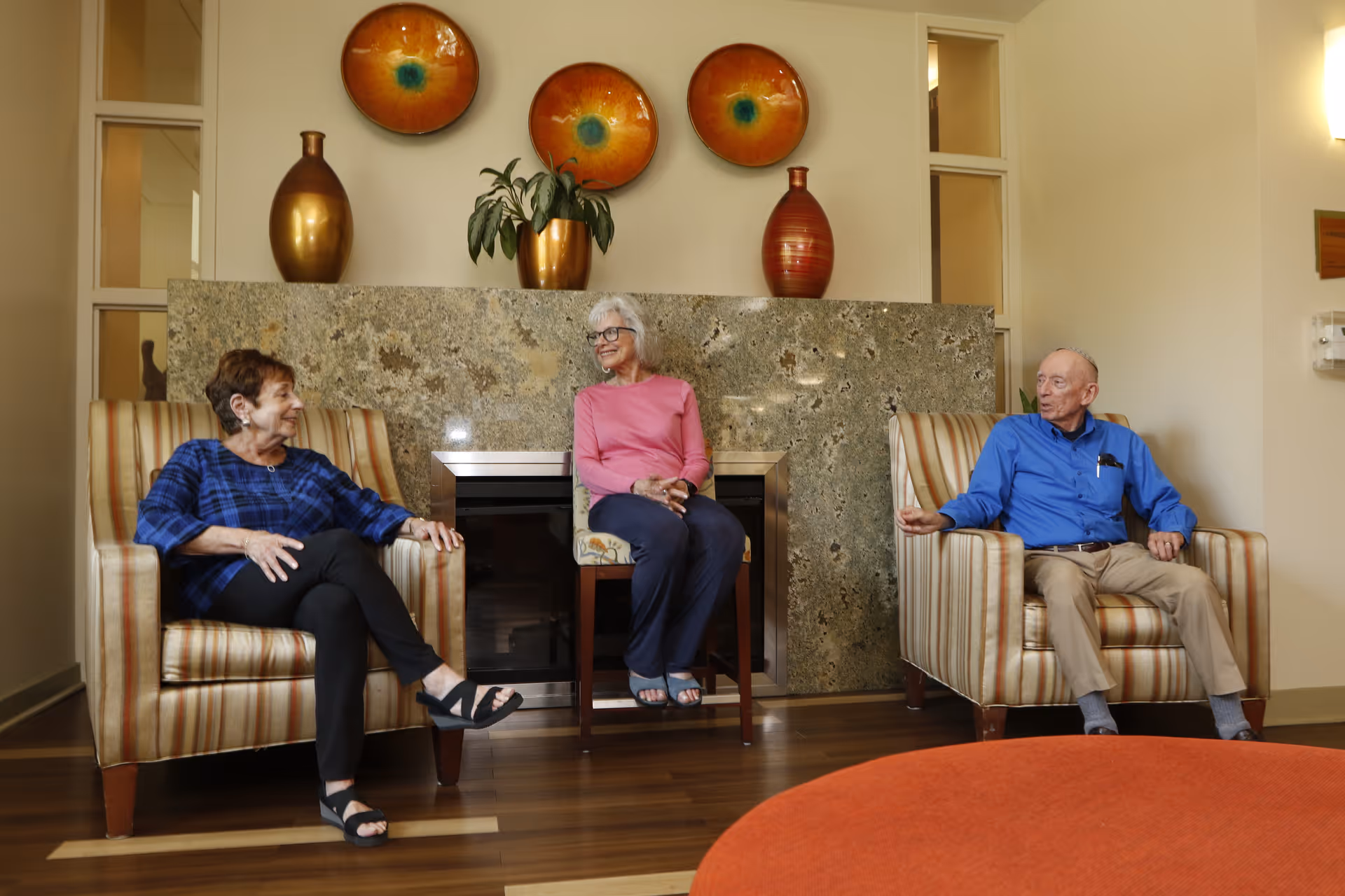 Three elderly people sitting and conversing in a living room area with two striped armchairs and a wooden chair in front of a stone fireplace. The room has decorative vases and plates on the mantel and a wooden floor.