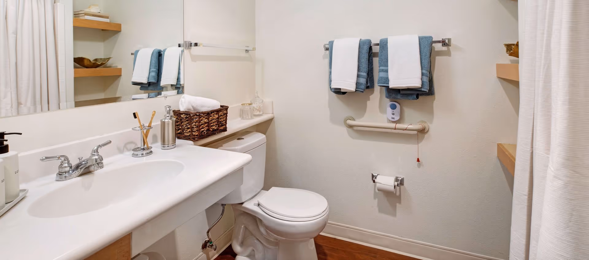 A clean, accessible bathroom with a sink and vanity, a toilet with a grab bar, a wall-mounted towel rack holding folded towels, mirror, shelving, and a shower curtain.