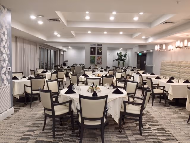 A spacious dining room with multiple round and rectangular tables covered with white tablecloths. Each table is set with black folded napkins, glassware, and small centerpieces. The room has modern lighting fixtures on the ceiling and walls, a patterned carpet, and large windows with curtains on one side.