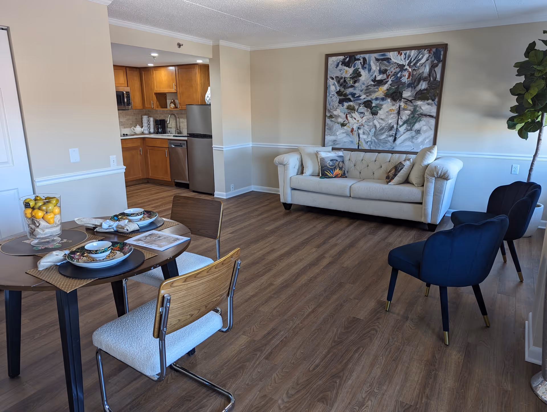 A bright and modern living area featuring a beige tufted sofa with decorative pillows, two dark blue chairs with gold-tipped legs, a large abstract painting on the wall, and a round dining table set for two with bowls, plates, and napkins. The room has wood flooring and an open view into a kitchen with wooden cabinets, a stainless steel refrigerator, and a dishwasher. A tall green plant is placed near the window.