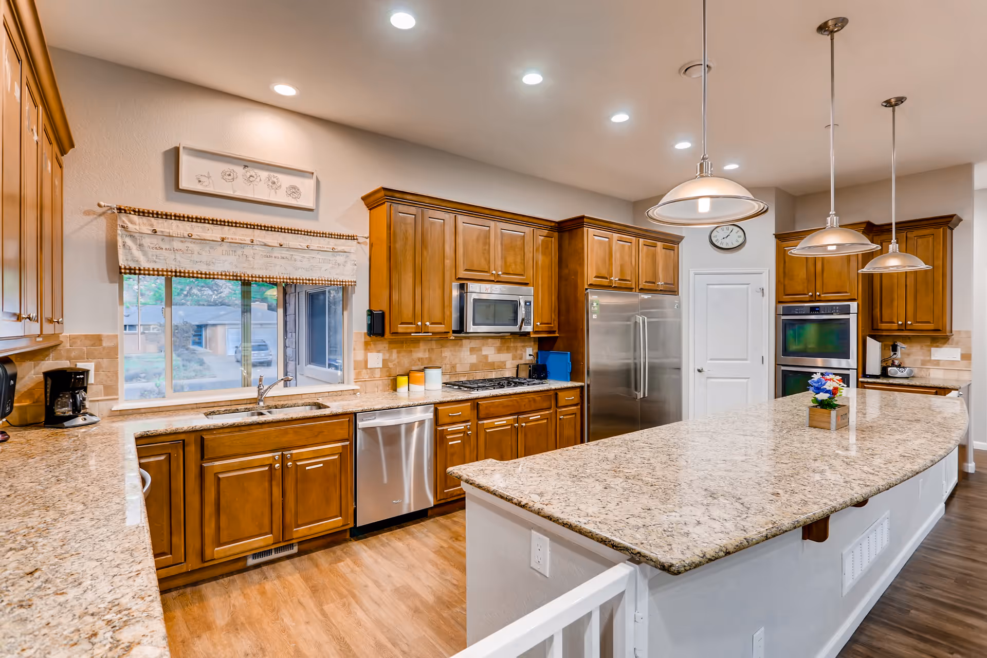 A spacious kitchen with wooden cabinets, granite countertops, and stainless steel appliances including a refrigerator, dishwasher, microwave, and double oven. There is a large kitchen island with pendant lights hanging above it and a small flower arrangement on the island. A window with a decorative valance overlooks the outside.