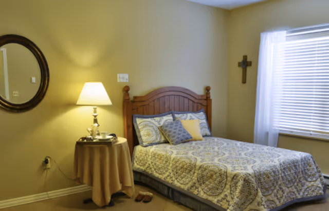 A neatly made bedroom with a wooden headboard, patterned bedspread, bedside table with a lamp and round mirror, and a window with blinds and a sheer curtain.