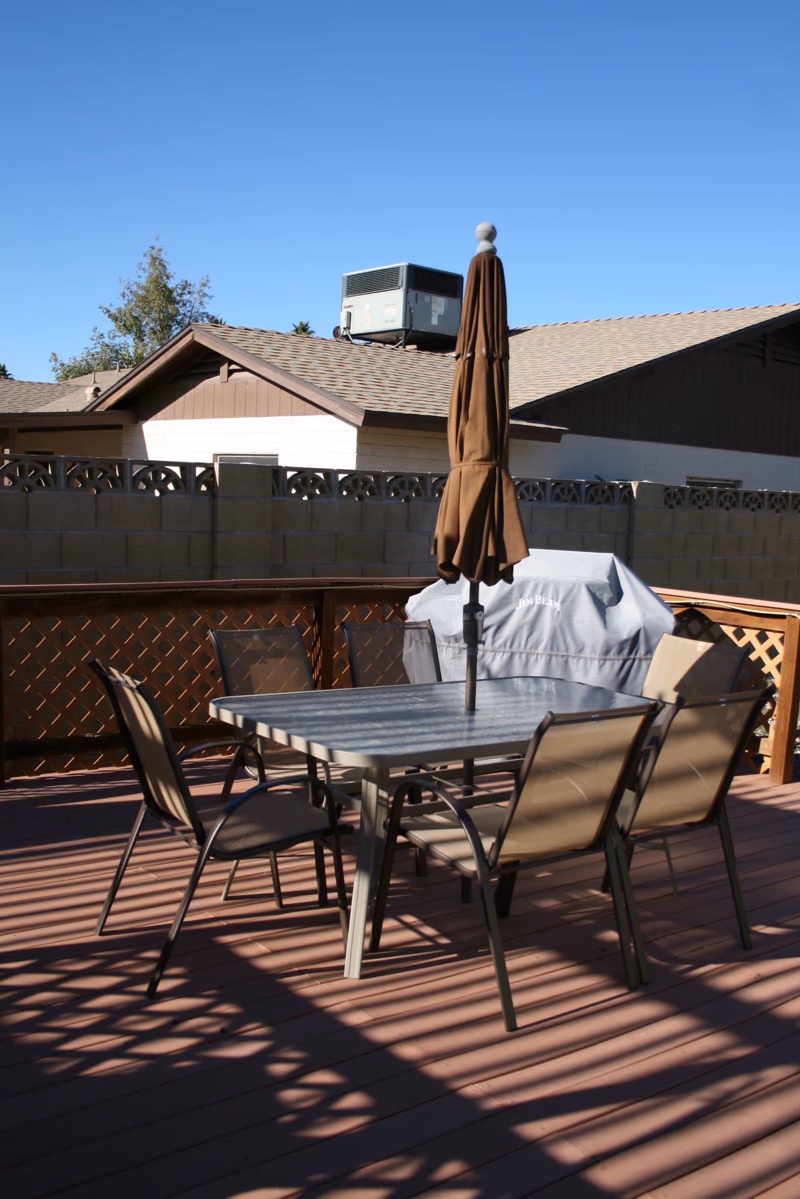 Wooden deck patio with a metal table, four chairs and a closed umbrella beside a covered grill, with a block wall and neighboring house under a clear blue sky.