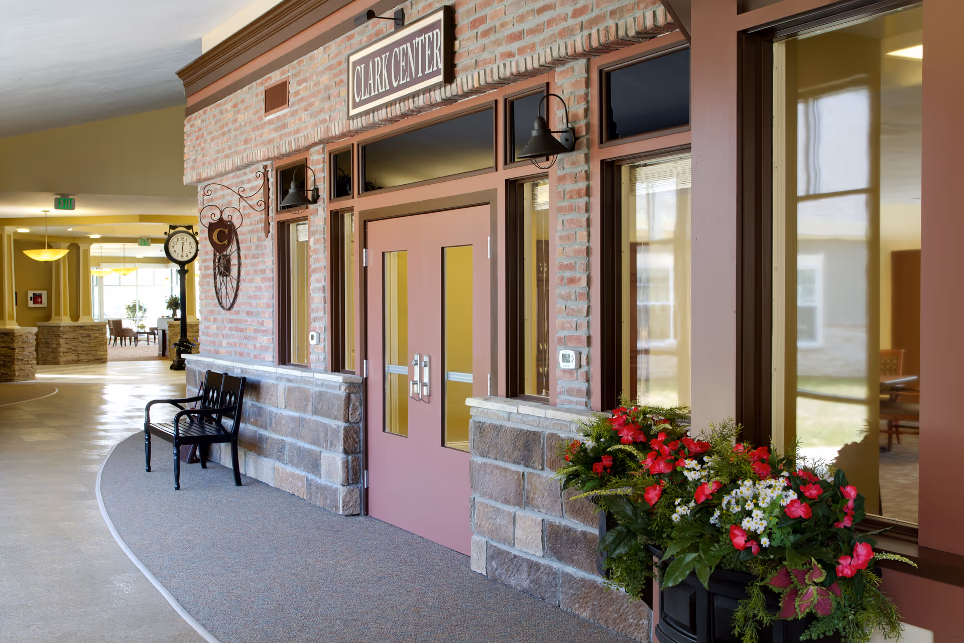 Interior hallway showing a brick-faced 'Clark Center' entrance with double doors, a bench, clock, and a planter of red flowers.