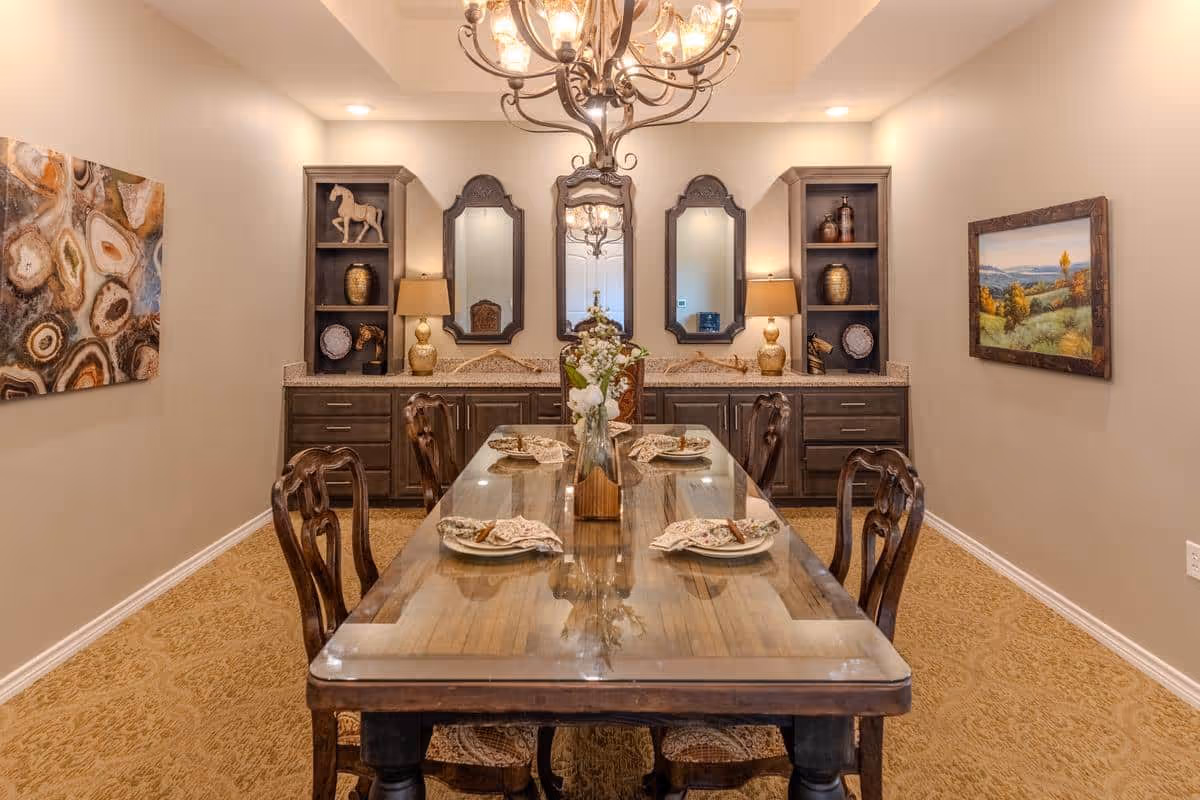 A formal dining room with a long wooden table set for six, featuring decorative plates and napkins. The room has beige walls, carpeted floor, and a chandelier hanging above the table. At the back, there are two mirrors flanked by built-in shelves and cabinets with lamps and decorative items. Artwork is displayed on the side walls.