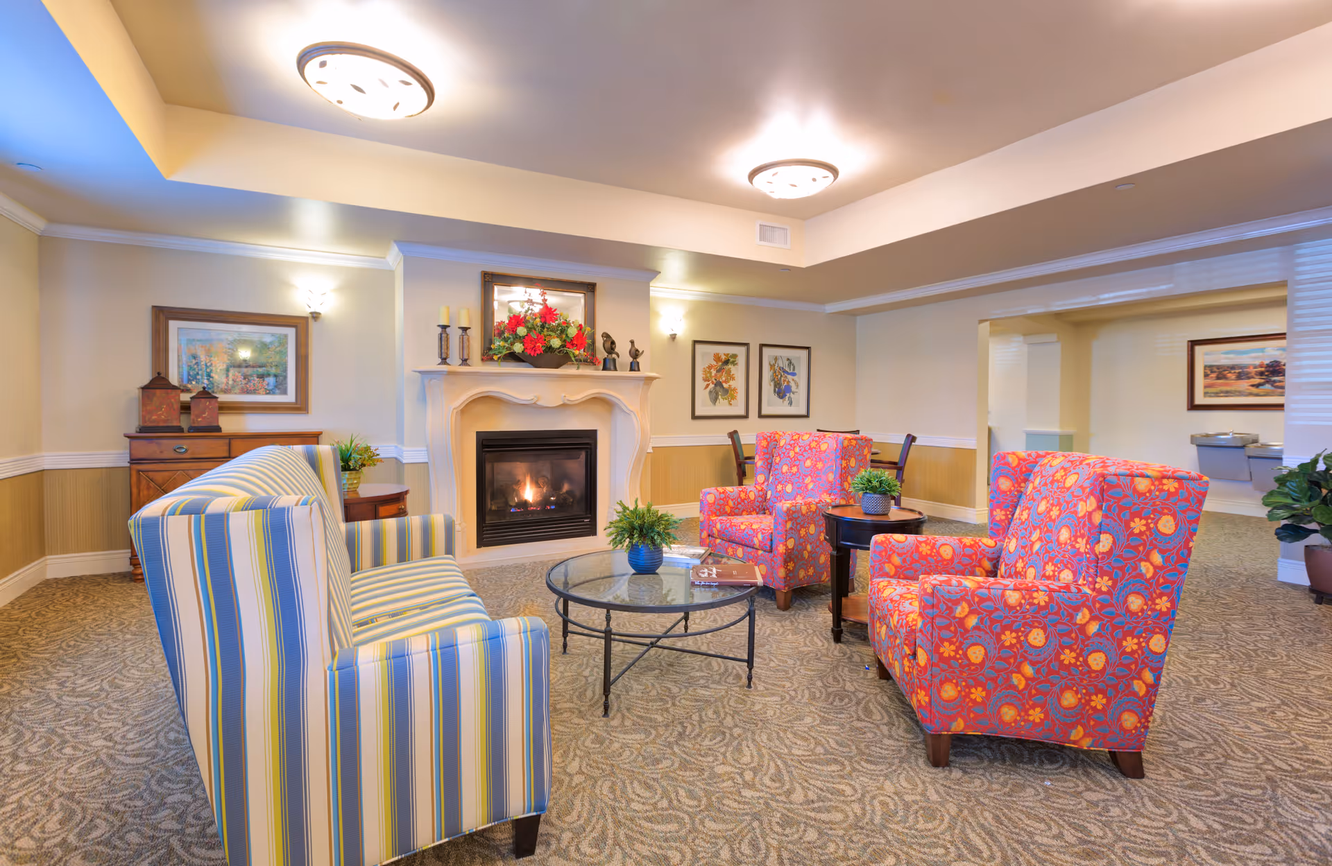 A cozy living room area in Shawnee Hills facility featuring a lit fireplace with decorative items on the mantel. The room has a striped sofa and two colorful patterned armchairs arranged around a glass coffee table with a small plant. The walls are adorned with framed artwork, and the space is warmly lit with ceiling and wall lights.