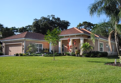 Single-story house with a red tile roof, beige exterior walls, a two-car garage, and a well-maintained green lawn with small trees and shrubs in front under a clear blue sky.