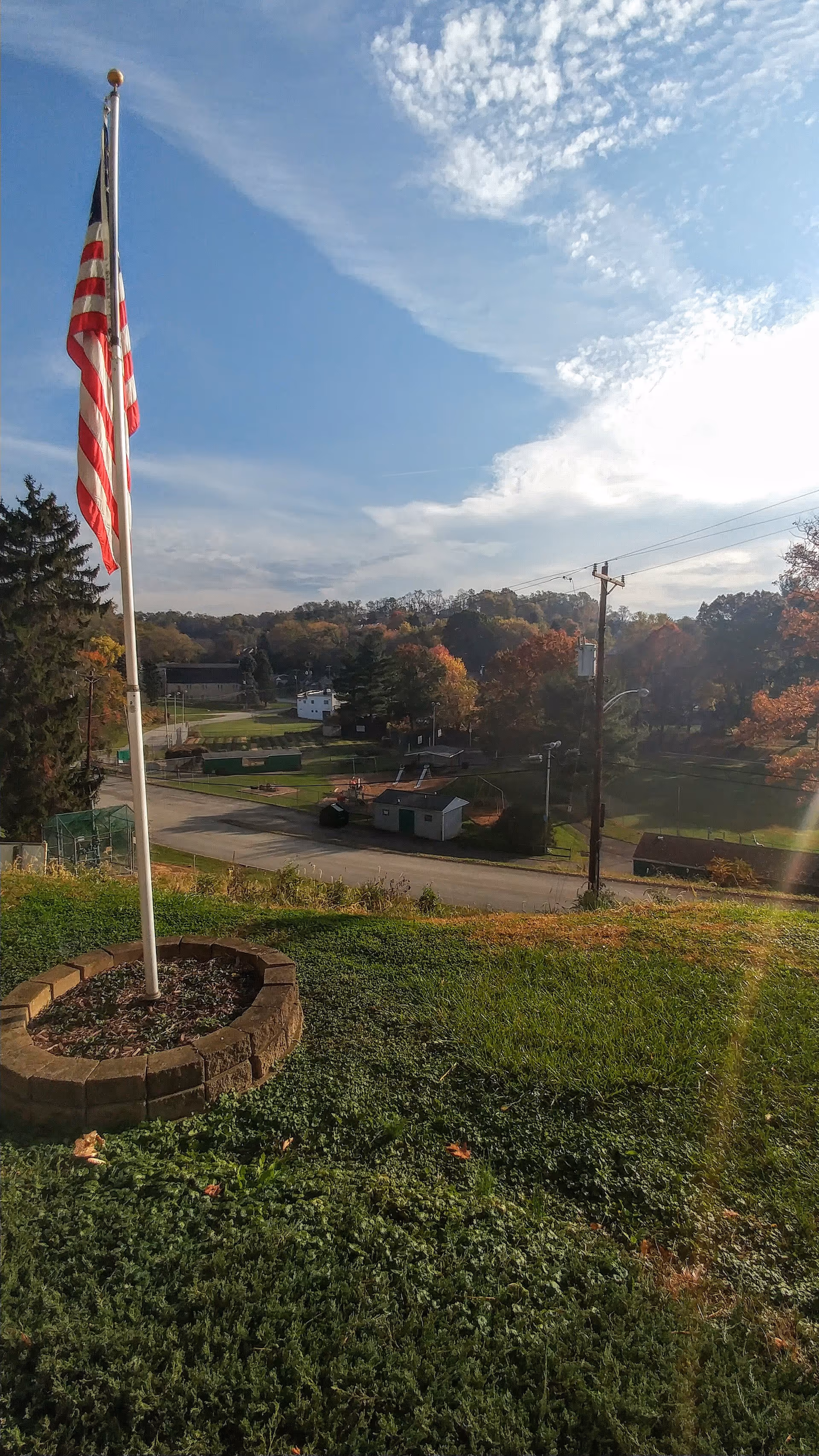 An outdoor scene featuring an American flag on a flagpole planted in a circular brick base surrounded by green grass. In the background, there is a road, several small buildings, trees with autumn foliage, and a partly cloudy blue sky.